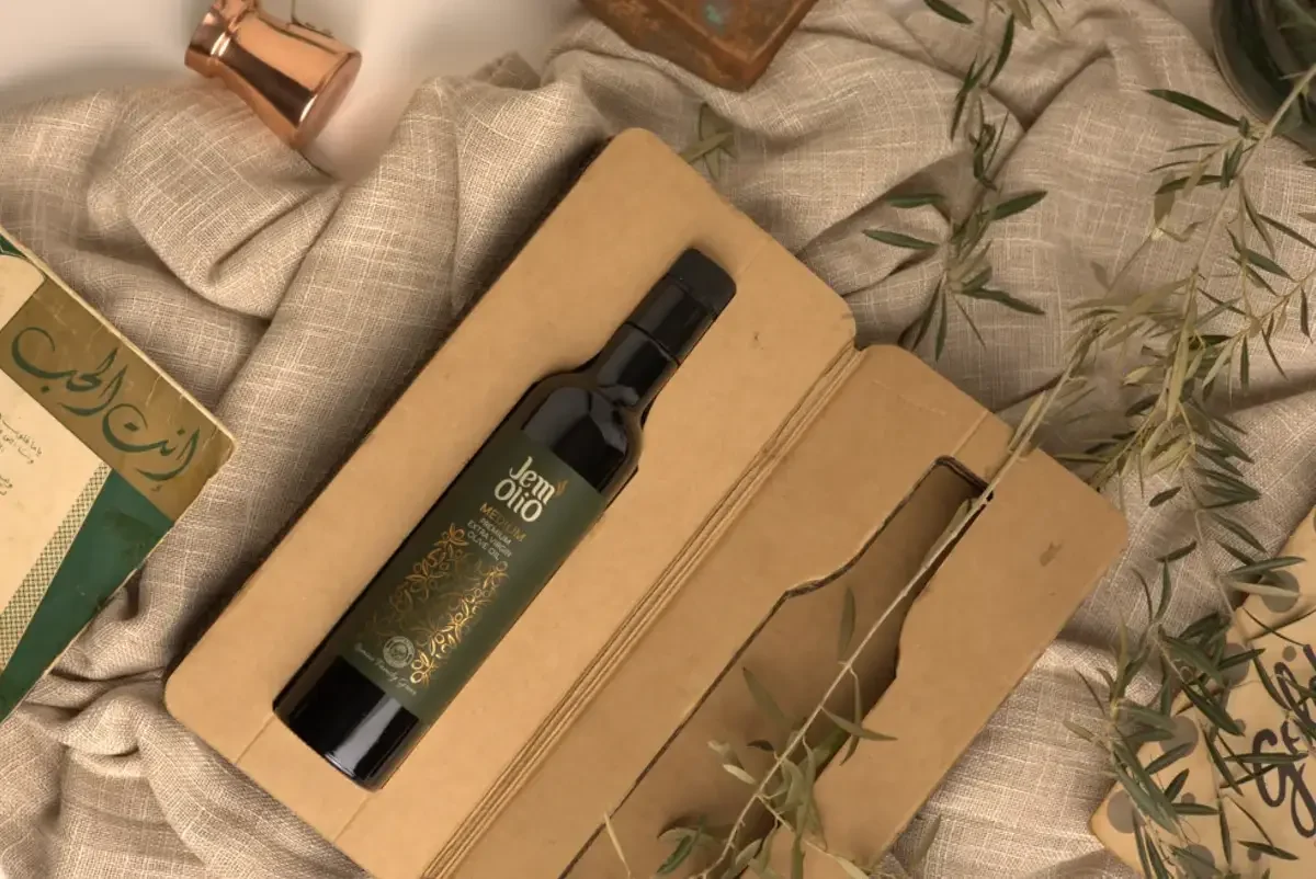 La bouteille Jemolio dans son coffret kraft, sur un lin clair, près d'un manuscrit et d'un pot de cuivre